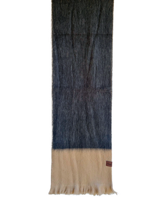 A long, narrow fuzzy scarf hanging from a wooden hanger against a plain white background. The scarf has a dark charcoal-to-black upper section and a warm beige lower section with fringe, plus a small brown leather tag near the bottom right.