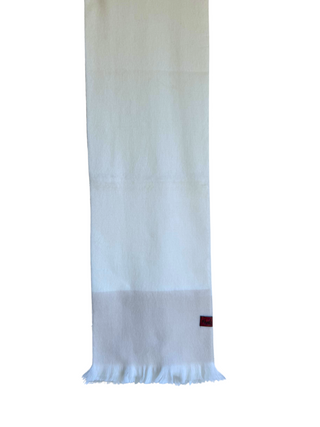 A white woven scarf with a fringed hem, hanging from a wooden hanger against a plain white background. A small dark red brand tag is visible near the lower right end.