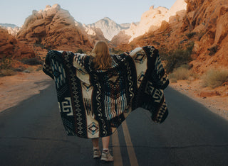 Person walking on a desert road with a patterned alpaca blanket, surrounded by rocky landscapes.